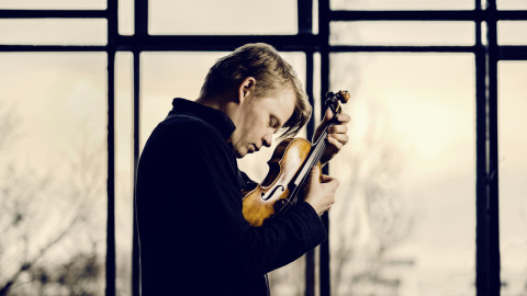 Mustapaitainen mies pitelee sylissään viulua lähellä päätään. Kuva Jyväskylä Sinfonia