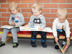 Kolme pientä lasta istuu penkillä tiiliseinän edessä ja katselee kuvakirjoja. Kuva Dolly Aittanen